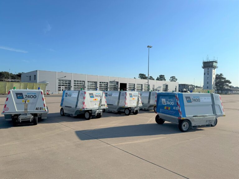 Cinq convertisseurs au sol à batterie itw gse 7400 sur l'aire de trafic avec un hangar, une tour de contrôle et un ciel bleu en arrière-plan.